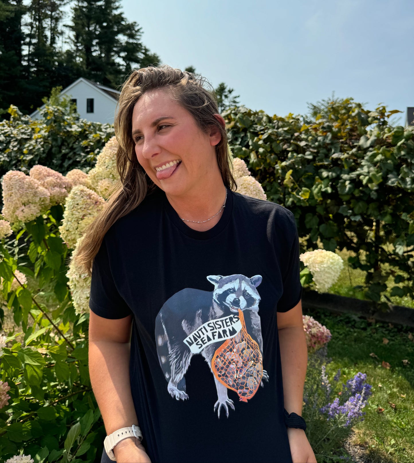 A person standing outdoors wearing a black t-shirt with a racoon design and the text 'Wild & Free'. There are flowers in the background.